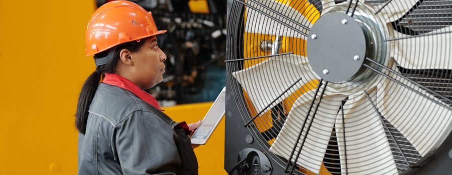 Young repair person in workwear and safety helmet checking air mover of huge industrial machine while working in factory